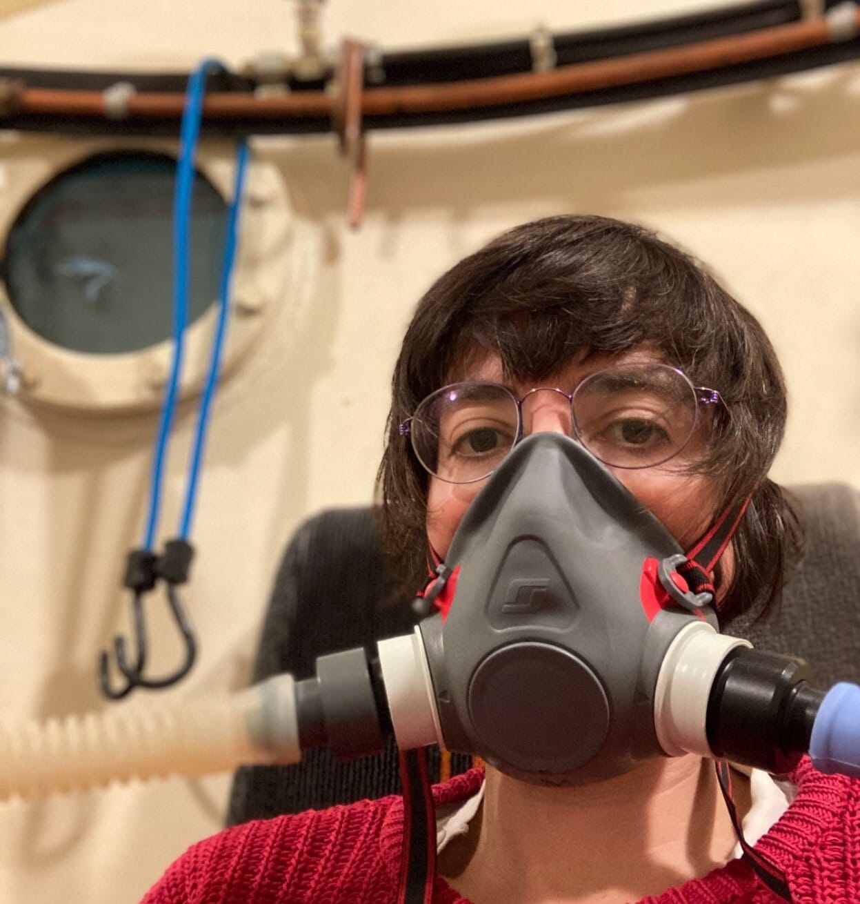 A white person with messy brown hair in a hyperbaric oxygen chamber w/ a nautical round window and a grey respirator on